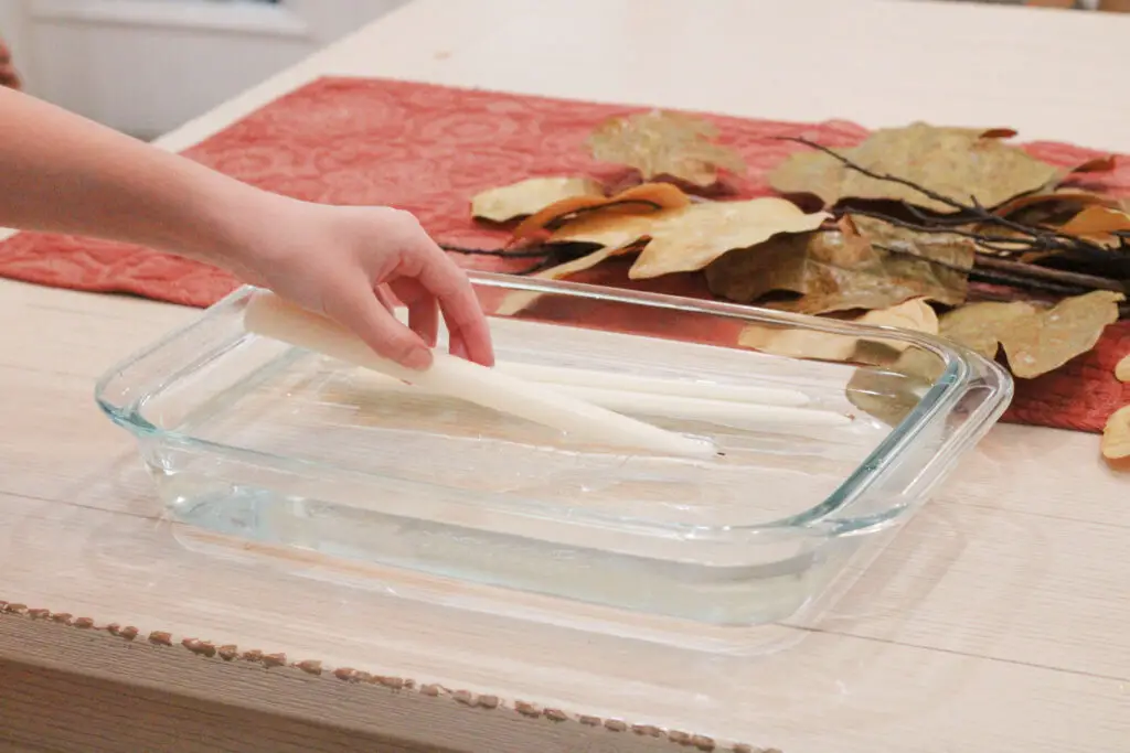 Close up of a hand putting a white candle into a glass baking dish with water in it. 