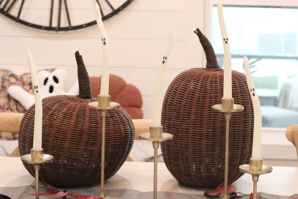 A close up of a dining table with gold candlestick holders with white tapered candles that have black ghosts faces on them, dark brown wicker pumpkins behind them, on a white dining table. 