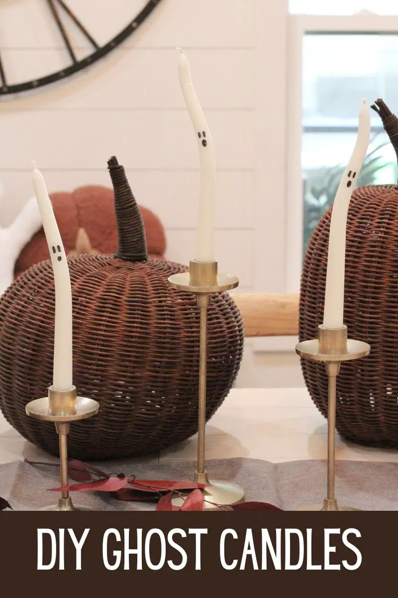 3 white tapered candles with black ghosts faces on them in gold candlestick holders, with brown wicker pumpkins behind them, on a white dining table with the text "DIY Ghost Candles" written at the bottom