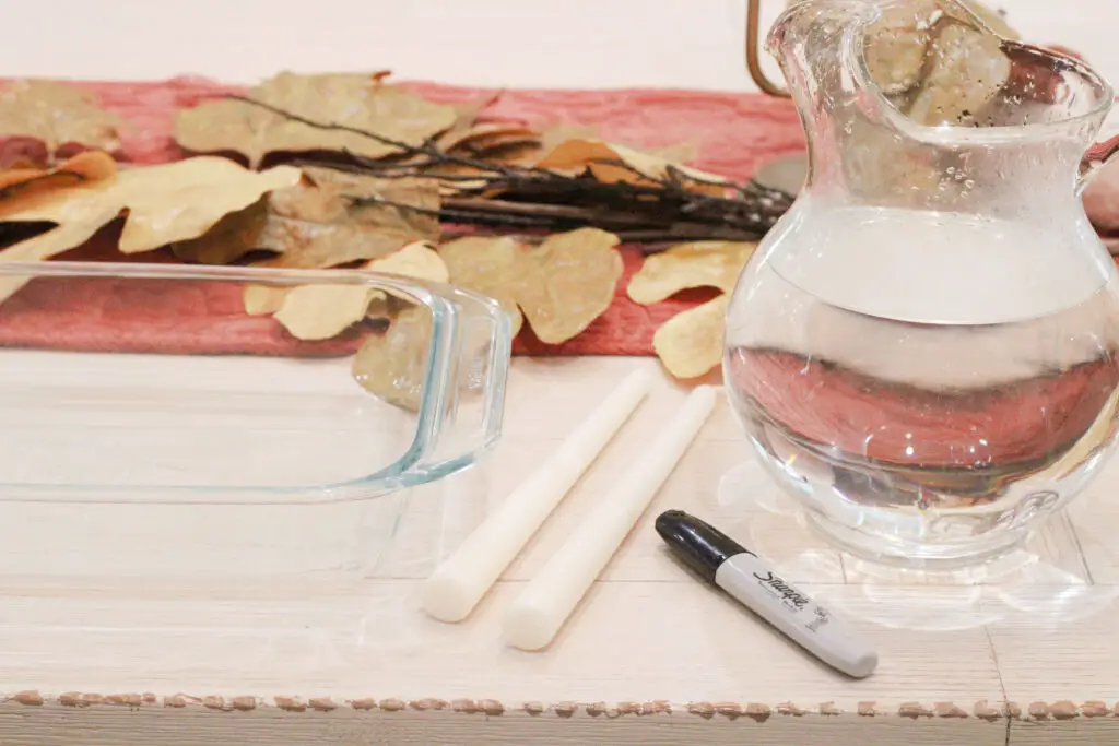 A close up of a glass container, 2 white tapered candlesticks, a black sharpie, and a glass pitcher with water in it. 