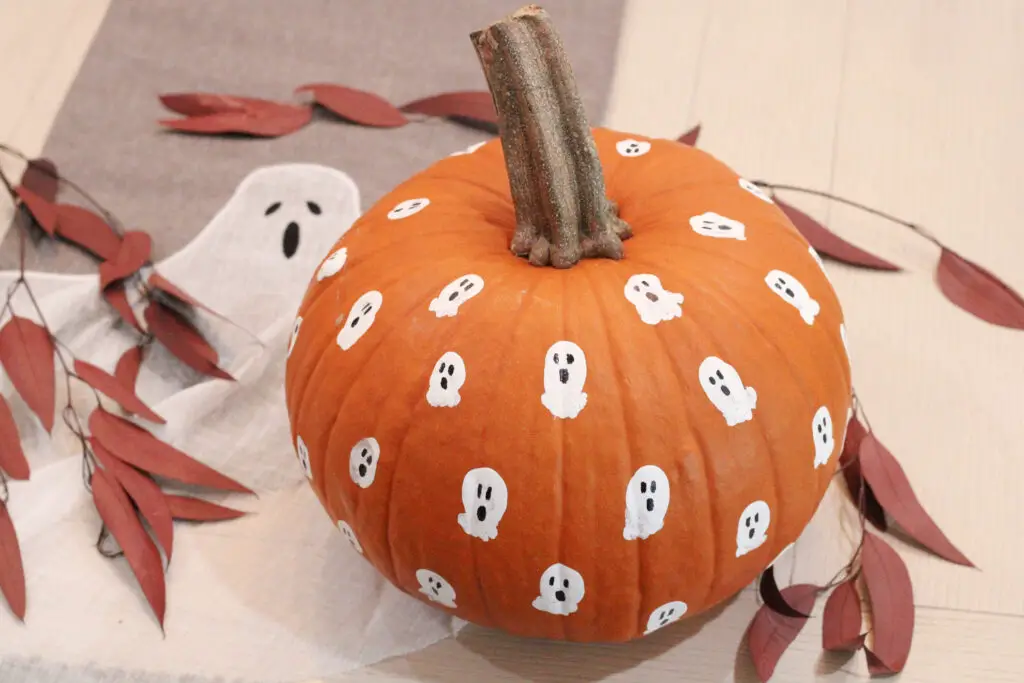 Close up of a real orange pumpkin with white tiny ghosts on it on a white table with red leaves around it and a ghost gray table runner