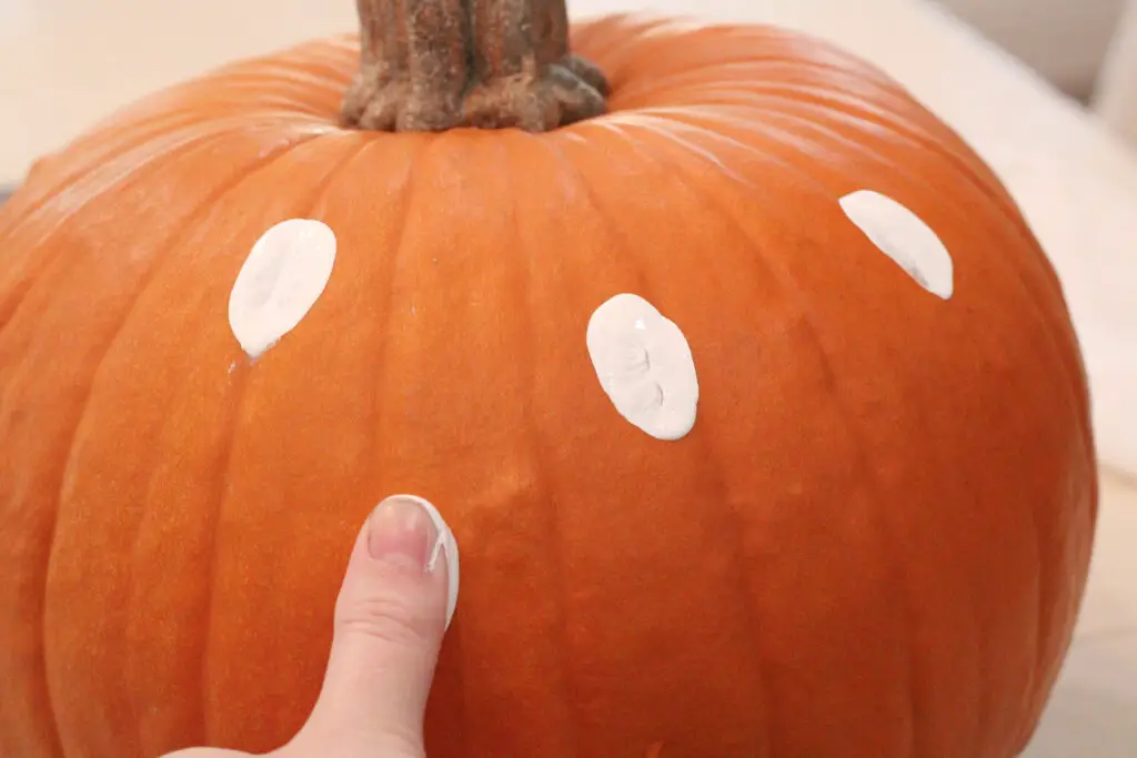 Close up of a real orange pumpkin with white dots of paint on it and a persons hand making a thumbprint onto the pumpkin with white paint