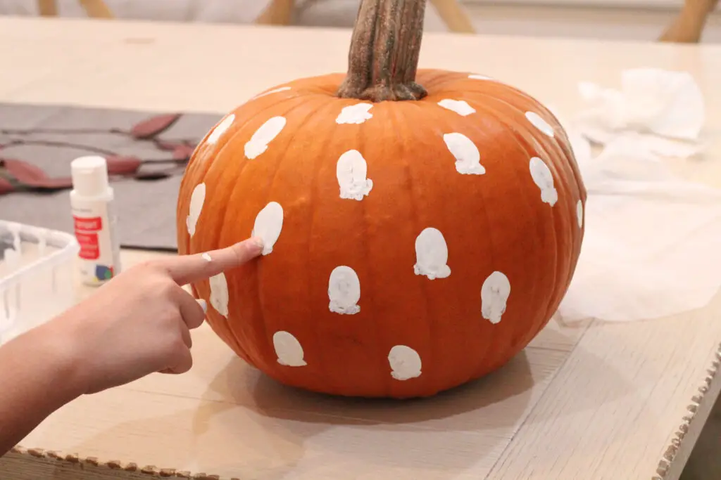 Close up of a real orange pumpkin, with a hand making fingerprints of white paint onto the pumpkin with white paint. 