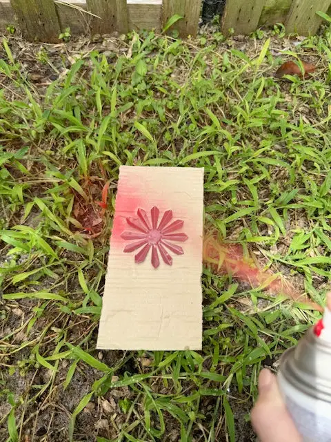 Close up of a piece of cardboard on the green grass with a red firework and a red spray paint can with paint coming out of it