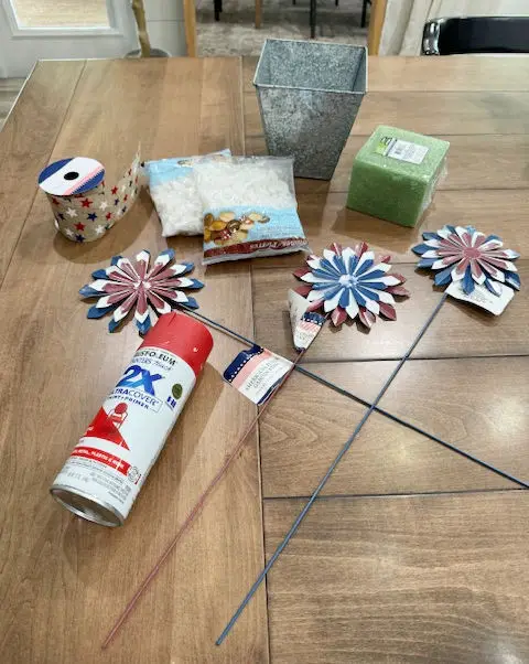 Close up of a red spray paint can, red white and blue ribbon with stars, white rocks, a silver vase, a green foam block, red white and blue firework garden stakes