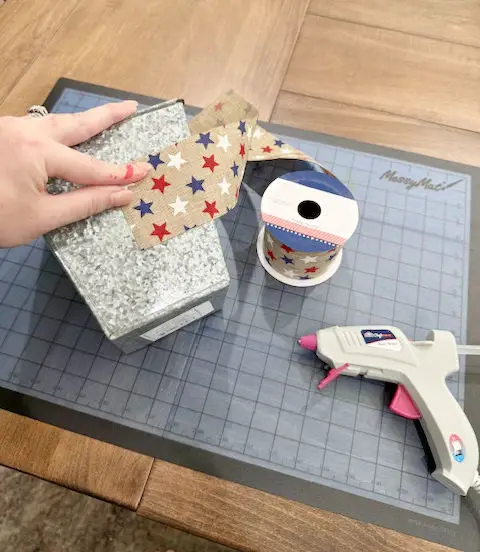 Close up of a silver vase with red white and blue ribbon and a hand with a hot glue gun