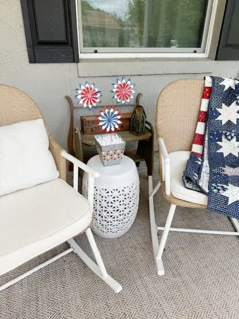 white and wicker rocking chairs next to a white table, on top of the table is red white and blue firework flowers in a silver vase, on a front porch