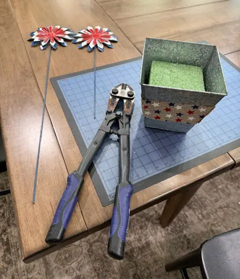 Close up of a wire cutter tool, with red white and blue patriotic firework garden stakes, one with longer stake then the other, silver vase with green foam floral block inside of it. 