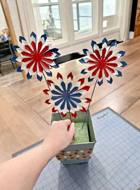 Close up of a hand on the red white and blue firework flowers placing them into the green foam block and silver vase