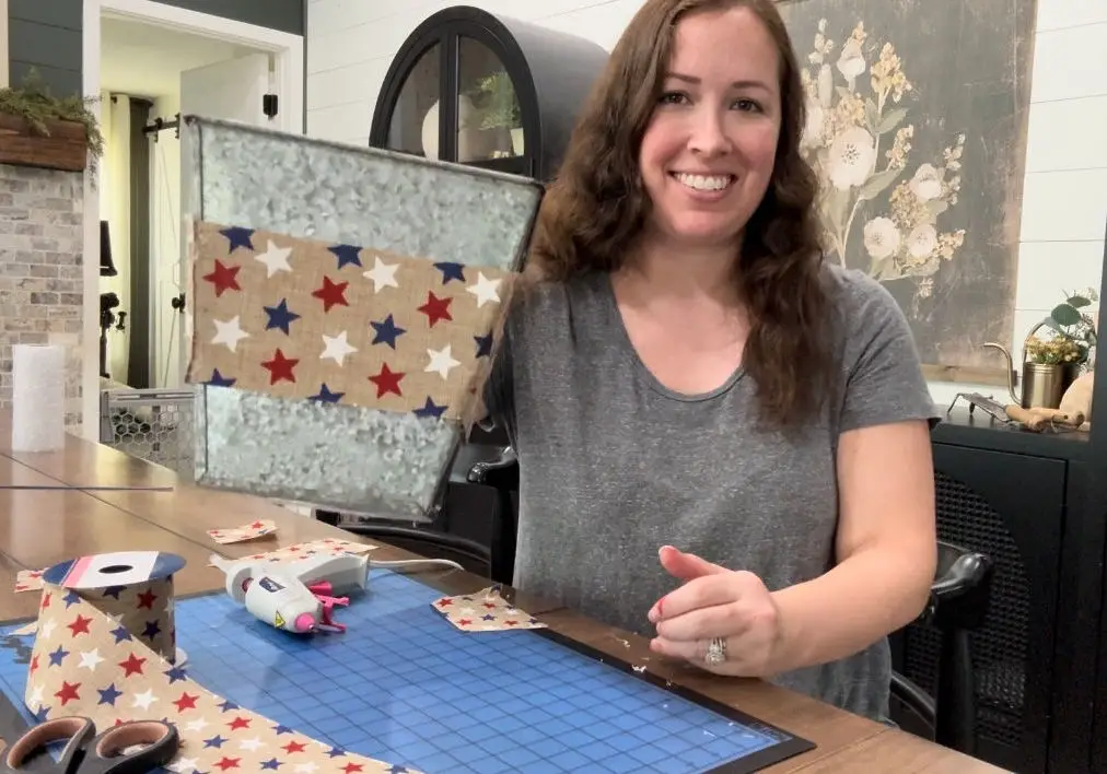 a woman with brown hair holding a silver vase with red white and blue patriotic ribbon with stars and a hot glue gun on a dark wood table and scissors